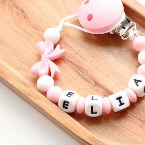 May include: Pink and white silicone baby teether with a clip and a bow. The teether has the name "Ellie" spelled out in black letters.