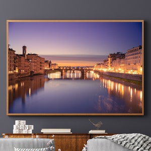 May include: A framed photograph of a cityscape at dusk with a bridge over a river. The buildings are lit up with warm yellow lights, and the water reflects the lights. The sky is a soft purple and pink.