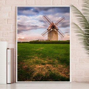 May include: A white framed photograph of a windmill in a field with a cloudy sky in the background.