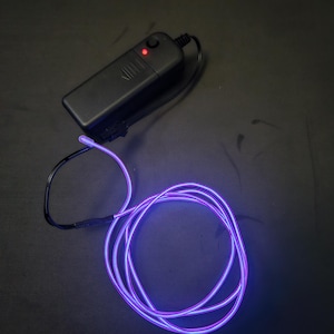 May include: A black battery pack with a red indicator light is connected to a coiled, glowing purple EL wire. The wire is a vibrant purple color and is designed for decorative lighting applications. The EL wire is coiled on a dark surface.