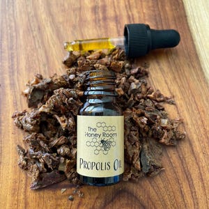 May include: A small brown glass bottle with a dropper filled with propolis oil. The bottle has a label that says "The Honey Room" and "Propolis Oil". The bottle is sitting on a wooden surface with a pile of brown propolis pieces around it.