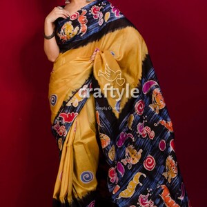 May include: A mustard yellow sari with a black border and colourful floral and abstract patterns. The sari has the text "craftyle" and "Simply Creative" printed on it. The sari is draped on a person.