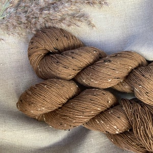 May include: Two skeins of brown yarn with black flecks. The yarn is twisted together and is lying on a light brown fabric background.