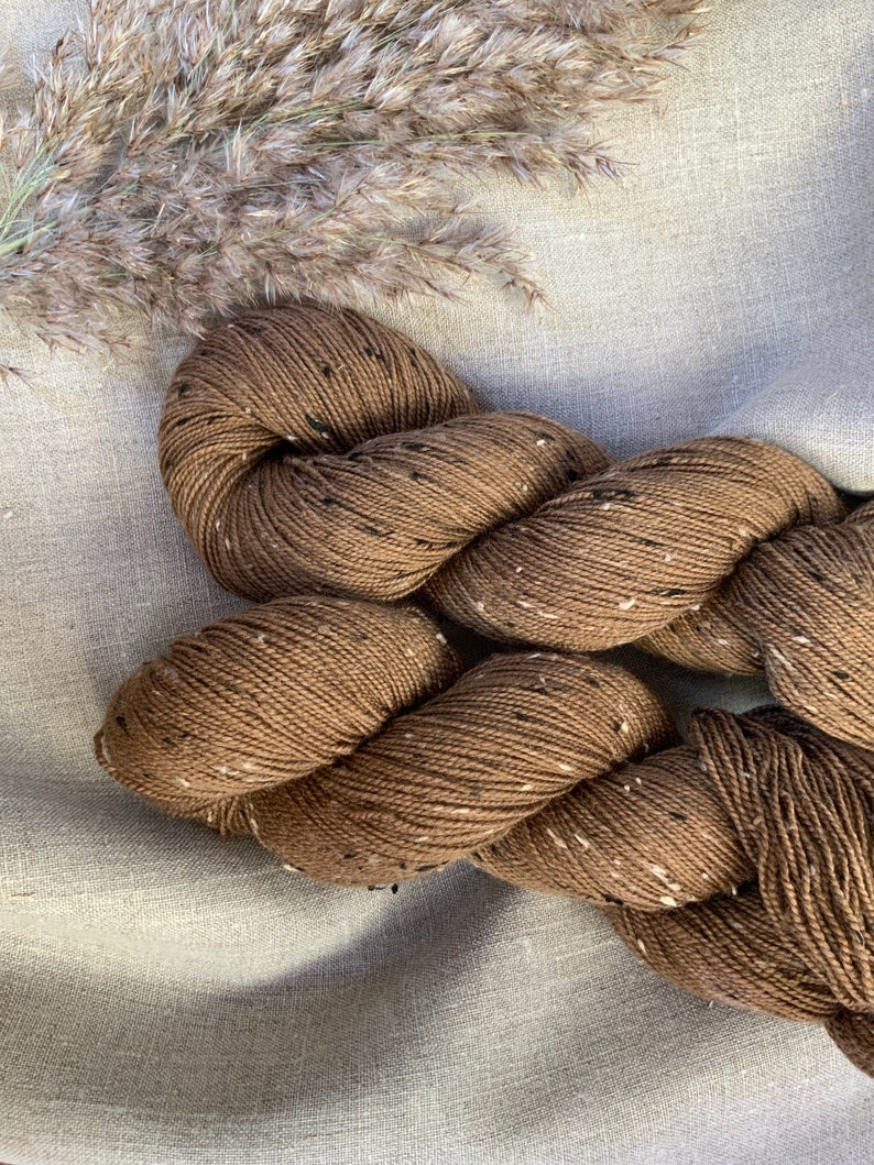 May include: Two skeins of brown yarn with black flecks. The yarn is twisted together and is lying on a light brown fabric background.