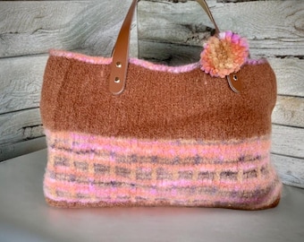 May include: A brown and pink tote bag with leather handles and a decorative pom-pom. The bag features a textured, wool-like material with a striped pattern in shades of pink and brown. The handles are a light brown.