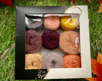 May include: A box of nine colorful yarn skeins in various shades of purple, peach, yellow, and orange. The yarn is arranged in a grid pattern, perfect for knitting or crochet projects. The box is black with a clear window.