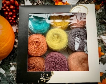 May include: A black and white box with a clear window, filled with nine skeins of colorful yarn. The yarn colors include teal, yellow, peach, gray, orange, purple, and brown. The box is decorated with gold floral accents.
