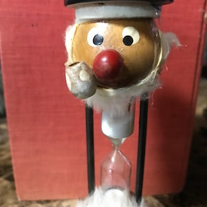 May include: A wooden hourglass with a whimsical face. The face has a black hat, white beard, brown eyes, a red nose, and a pipe. The hourglass is made of glass and metal.