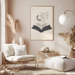 以下が含まれることがあります： 開いた本、三日月、星の白黒イラストが描かれた額入りプリント。このアートワークは、モダンな椅子とオットマンを備えたニュートラルな色調の部屋に展示されています。
