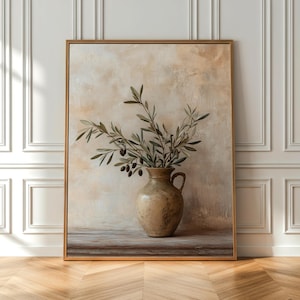 Op de afbeelding: Een ingelijste wandkunstprint met een gestructureerde, beige keramische kan met één handvat. In de kan staat een tak met groene olijfbladeren en kleine donkere olijven tegen een zachte, gemêleerde crème en beige achtergrond.