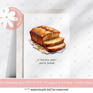May include: A greeting card with a watercolor illustration of a loaf of banana bread sliced and drizzled with honey. The text on the card reads "in banana years you're bread!"