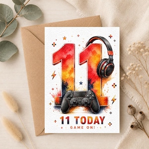 Può includere: Un biglietto d'auguri rettangolare con il numero 11 in un gradiente acquerello rosso, arancione e giallo. Un controller di gioco nero si trova alla base dei numeri e delle cuffie nere poggiano sul secondo 1. Il testo recita 11 TODAY GAME ON.