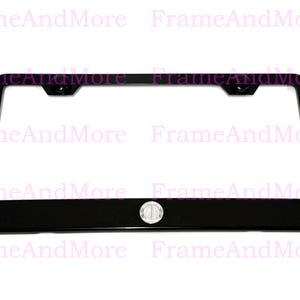 May include: A black license plate frame with a rectangular shape. The frame has a glossy finish and features a small, circular emblem at the bottom center. The frame is designed to hold a standard license plate.