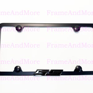 May include: Black license plate frame with rounded corners and four decorative black ball accents. The frame features the letters "SS" in the center. The text "FrameAndMore" is visible in the background.
