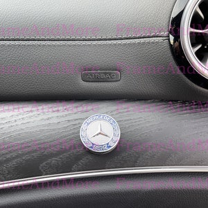May include: Close-up of a gray car interior with a wood-grain trim. The trim has a silver Mercedes-Benz emblem with a blue background. The text "AIRBAG" is visible on the dashboard.