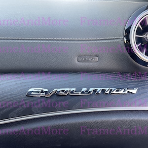May include: A close-up of a black car interior with a silver "Evolution" emblem on a wood-grain panel. The panel is located below a black leather dashboard with a silver air vent.