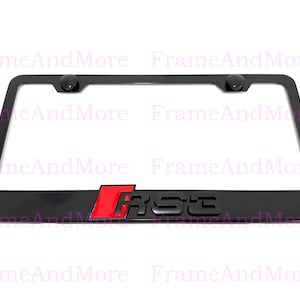 May include: Black license plate frame with the letters "RS3" in a red and black design. The frame has two black screw covers at the top. The frame is rectangular and has rounded corners.