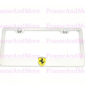 May include: A rectangular silver-colored metal license plate frame featuring a centered shield emblem with a black prancing horse on a yellow background. The frame includes two circular mounting points at the top for secure attachment.