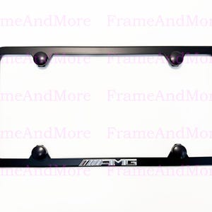 May include: Black license plate frame with four rounded mounting points. The frame has the AMG logo in silver and black at the bottom. The frame is rectangular and has rounded corners.