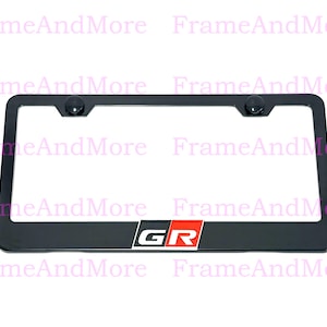 May include: A rectangular black license plate frame featuring a centered GR logo with white lettering on a black background and a red square accent. The frame includes two circular black mounting points at the top corners for secure attachment.