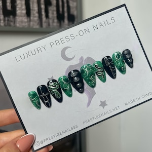 May include: Set of ten green and black press-on nails with snake and glitter designs. The nails are displayed on a white card with the text "LUXURY PRESS-ON NAILS" and "@PRESTIGENAILSSS PRESTIGENAILS.NET MADE IN CAMD".