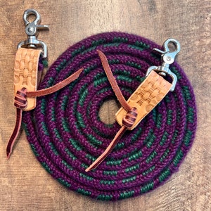 May include: A coiled purple and green rope with two leather and metal attachments. The leather attachments have a woven design and are connected to a silver-colored metal clasp. The rope is on a wooden surface.