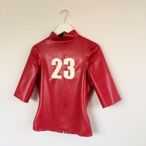 May include: A red latex top with a high collar and elbow-length sleeves. The front of the garment features the number 23 in large white lettering. The material has a smooth, glossy finish and is displayed on a white hanger against a plain wall.