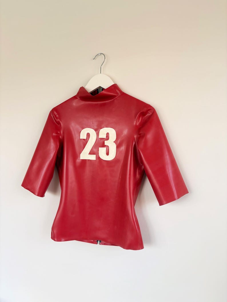 May include: A red latex top with a high collar and elbow-length sleeves. The front of the garment features the number 23 in large white lettering. The material has a smooth, glossy finish and is displayed on a white hanger against a plain wall.