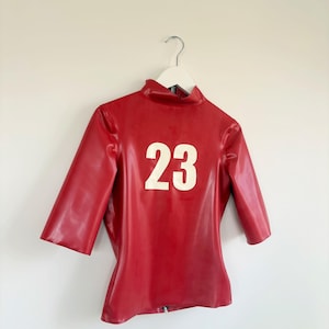 Op de afbeelding: Een rood latex shirt met een hoge kraag en ellebooglange mouwen. Op de voorzijde staat het getal 23 in grote witte blokletters. Het kledingstuk heeft een gladde, glanzende afwerking en een zichtbare ritssluiting aan de onderkant.