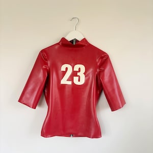 May include: A red latex top with half-length sleeves and a high collar. The garment features the number 23 printed in large white numerals on the front centre. It is displayed on a white hanger and includes a visible zip at the bottom hem.