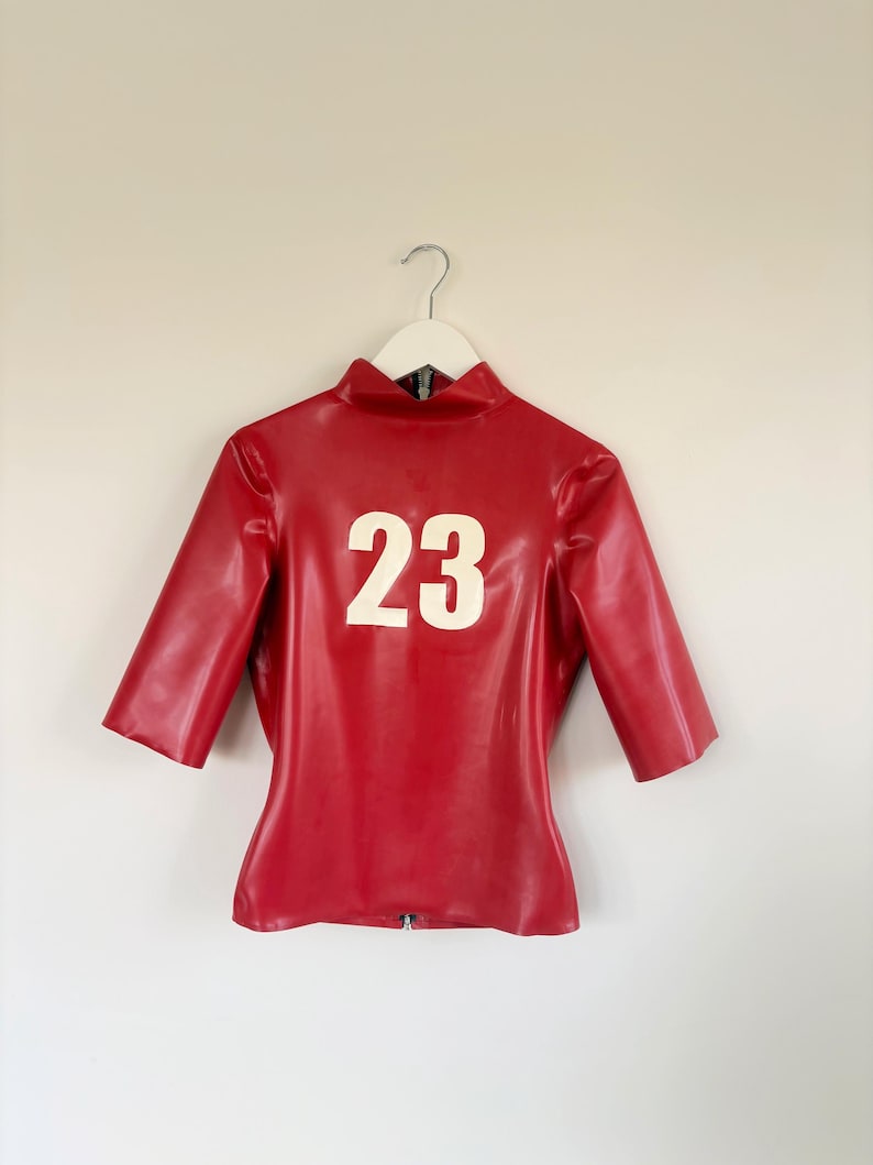 May include: A red latex top with half-length sleeves and a high collar. The garment features the number 23 printed in large white numerals on the front centre. It is displayed on a white hanger and includes a visible zip at the bottom hem.