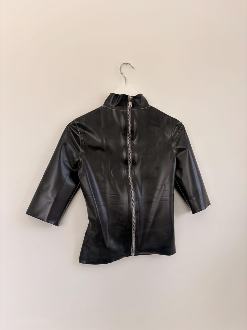 May include: A black, shiny, short-sleeved top with a high mock neck collar. The garment features a full-length silver-toned metal zip running vertically down the centre front, extending from the collar to the bottom hem of the shirt.