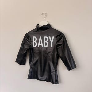 May include: A black mock neck top with three-quarter length sleeves and a shiny finish. The word BABY is printed in large white capital letters across the centre of the chest. The garment is displayed on a white hanger against a plain wall.