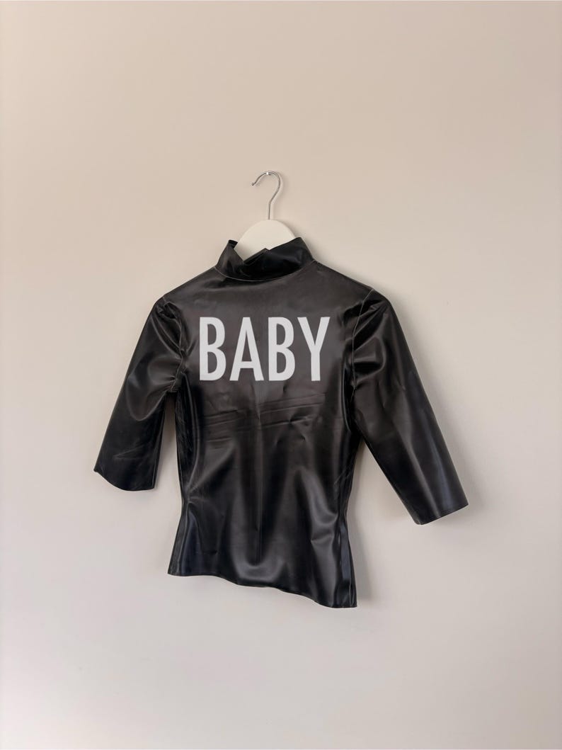 May include: A black mock neck top with three-quarter length sleeves and a shiny finish. The word BABY is printed in large white capital letters across the centre of the chest. The garment is displayed on a white hanger against a plain wall.