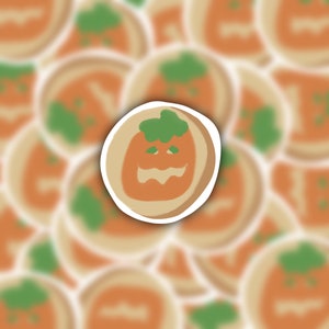 May include: A close-up of a pumpkin-shaped cookie with a green stem and a smiling jack-o'-lantern face. The cookie is surrounded by other blurred pumpkin cookies.