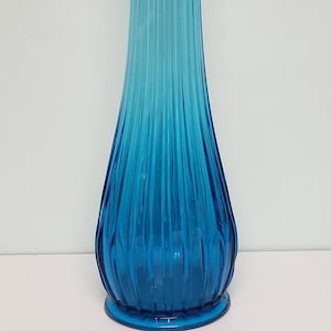 May include: A tall, blue glass vase with a flared opening and a ribbed design. The vase has a gradient of color, with a lighter blue at the top and a darker blue at the bottom.