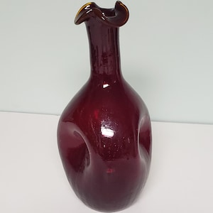 May include: A deep red glass vase with a unique, organic shape. The vase has a flared, ruffled rim with a golden-brown edge. The body of the vase has a bulbous form with a smooth, glossy finish, creating a striking decorative piece.