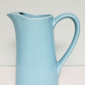 May include: A light blue ceramic pitcher with a wide mouth and a handle. The pitcher is tall and slender.