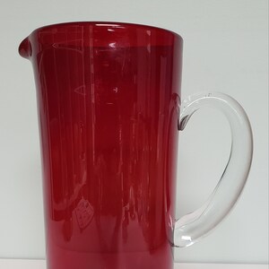 May include: A red glass pitcher with a clear glass handle. The pitcher is cylindrical in shape and has a wide mouth.