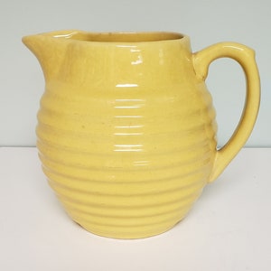 May include: A yellow ceramic pitcher with a ribbed design. The pitcher has a wide mouth and a handle.