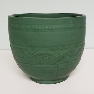 May include: A green ceramic planter with a decorative embossed design around the middle. The planter has a rounded bottom and a slightly flared top.