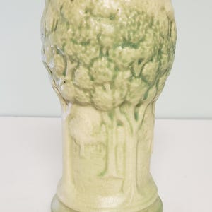 May include: A light green ceramic vase with a textured tree design. The vase has a cylindrical shape with a wide opening and a detailed relief of a tree and foliage. The base is a darker shade of green.