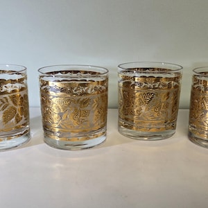 May include: Four clear drinking glasses with a gold floral design. The glasses have a gold band around the top and bottom, and a detailed gold pattern in the center. The glasses are cylindrical and sit on a white surface.