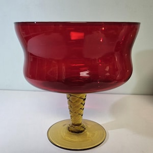 May include: A ruby red glass bowl with a wide, flared rim sits atop a twisted, golden stem and a circular base. The bowl's rich color and elegant design suggest it is a decorative piece, possibly for serving or display.