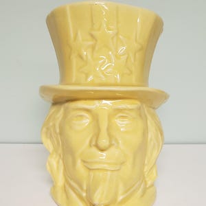 May include: A yellow ceramic planter shaped like a person's head wearing a hat with stars on it.