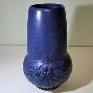 May include: A cobalt blue ceramic vase with a bulbous base and a cylindrical top. The base features a raised relief design of leaves and berries. The vase has a smooth, matte finish and is suitable for displaying flowers or as a decorative item.