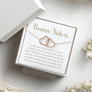 May include: A rose gold necklace with two interlocking heart pendants, presented in a white gift box. The card inside reads "Bonus Sisters" with a heartfelt message about family and appreciation. Delicate white flowers and fabric surround the box.