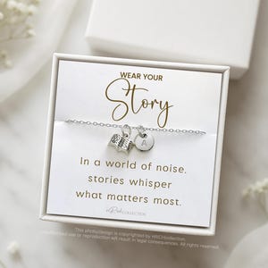 Może przedstawiać: Srebrna bransoletka z zawieszką w kształcie książki i okrągłą zawieszką z literą "A". Na karcie napisano "WEAR YOUR Story" i "In a world of noise, stories whisper what matters most."