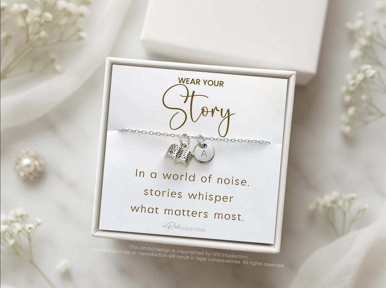 Może przedstawiać: Srebrna bransoletka z zawieszką w kształcie książki i okrągłą zawieszką z literą "A". Na karcie napisano "WEAR YOUR Story" i "In a world of noise, stories whisper what matters most."