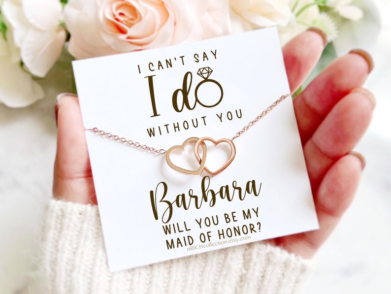 Peut inclure: Une carte blanche avec le texte "Je ne peux pas dire oui sans toi" et "Veux-tu &ecirc;tre ma demoiselle d'honneur ?" avec un collier en or rose avec deux c&oelig;urs entrelac&eacute;s. La carte comprend &eacute;galement le nom "Barbara" et le site Web "nRICHcollection.etsy.com".